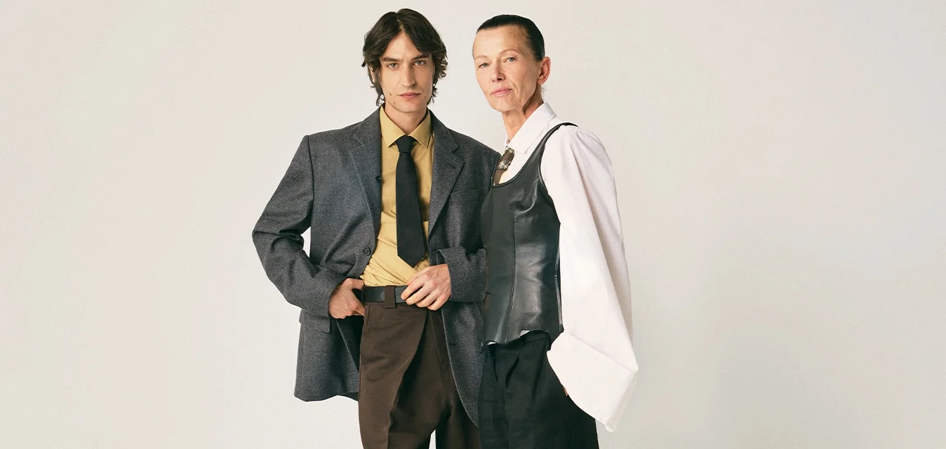 Due adulti si trovano di fronte a uno sfondo semplice indossando abbigliamento formale elegante: uno in un blazer grigio e cravatta, l'altro in una camicia bianca con un gilet nero.