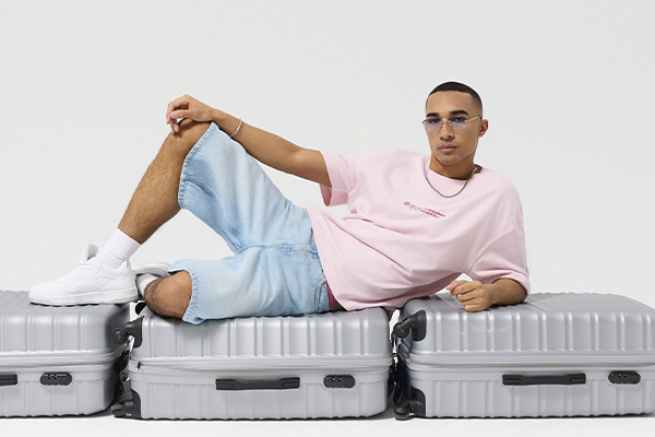 Modello con T-shirt rosa e shorts in denim sdraiato su valigie argento.