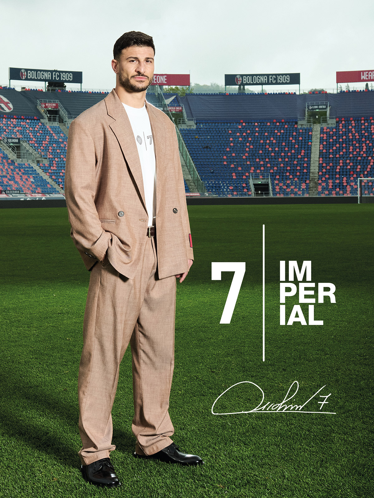 Man in beige suit standing on a football pitch inside a stadium with seating and a bold '7 Imperial' graphic