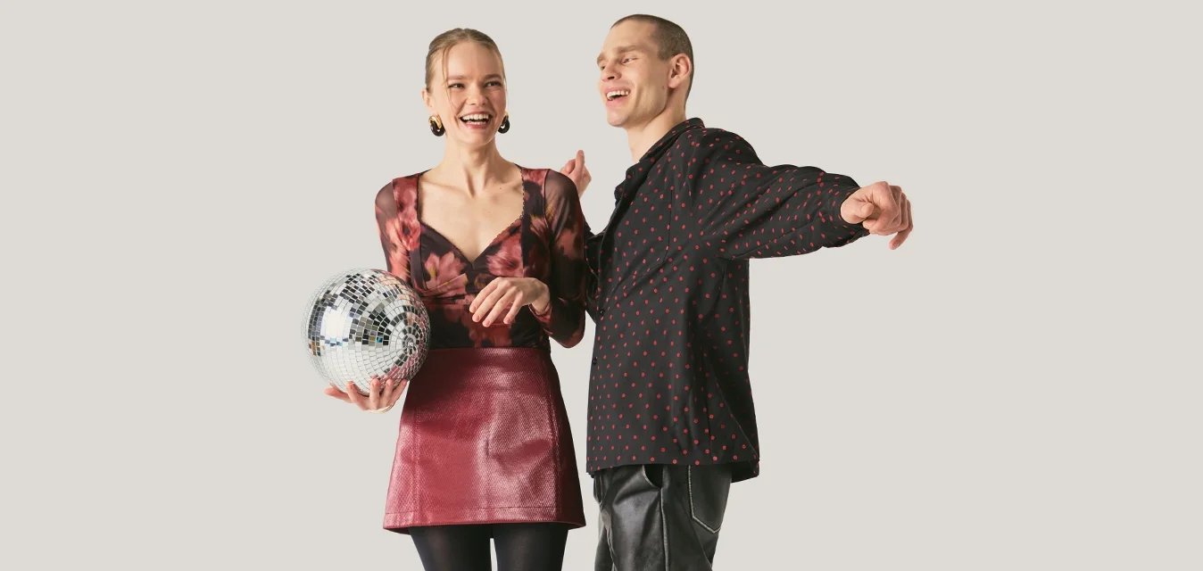 Donna che tiene una palla da discoteca, indossa una maglietta floreale e una gonna rossa, ride con un uomo in camicia nera a motivo e pantaloni di pelle che ballano.
