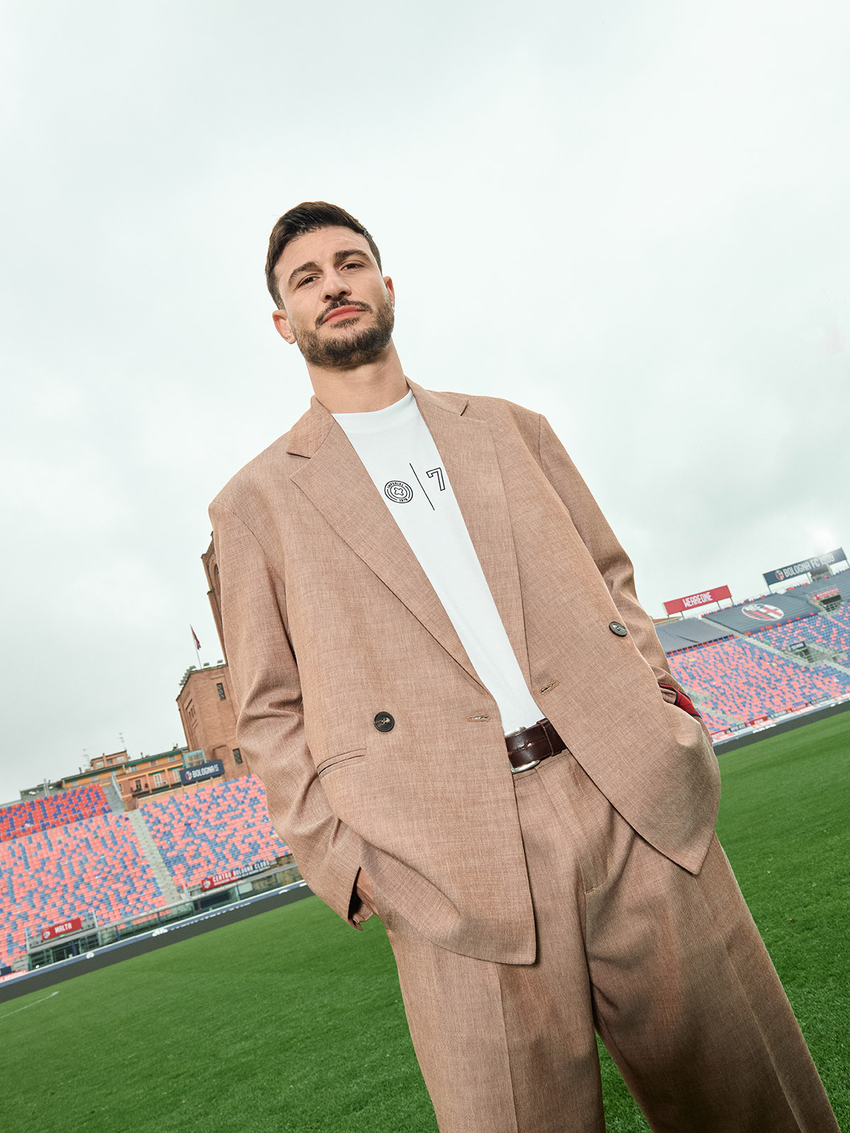 Man in beige suit with hands in pockets standing on a football stadium pitch with colourful stands and cloudy sky