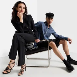  due modelle posano in un ambiente minimalista. Una indossa un completo nero e tacchi neri con cinturini; l'altra sfoggia una camicia blu e pantaloni corti neri, seduta.