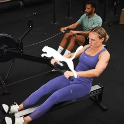 Macchina da remare impostata in una palestra. Due utenti in abbigliamento sportivo; uno indossa leggings viola e una canottiera, l'altro una maglietta verde. Pavimentazione nera e attrezzatura.