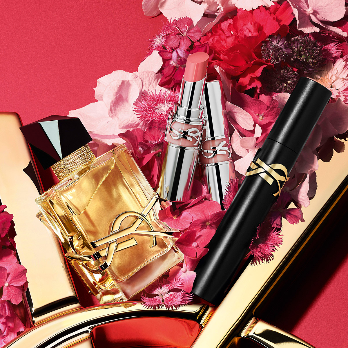 Un profumo Libre, due rossetti e un mascara YSL sono elegantemente disposti tra vivaci fiori rosa.