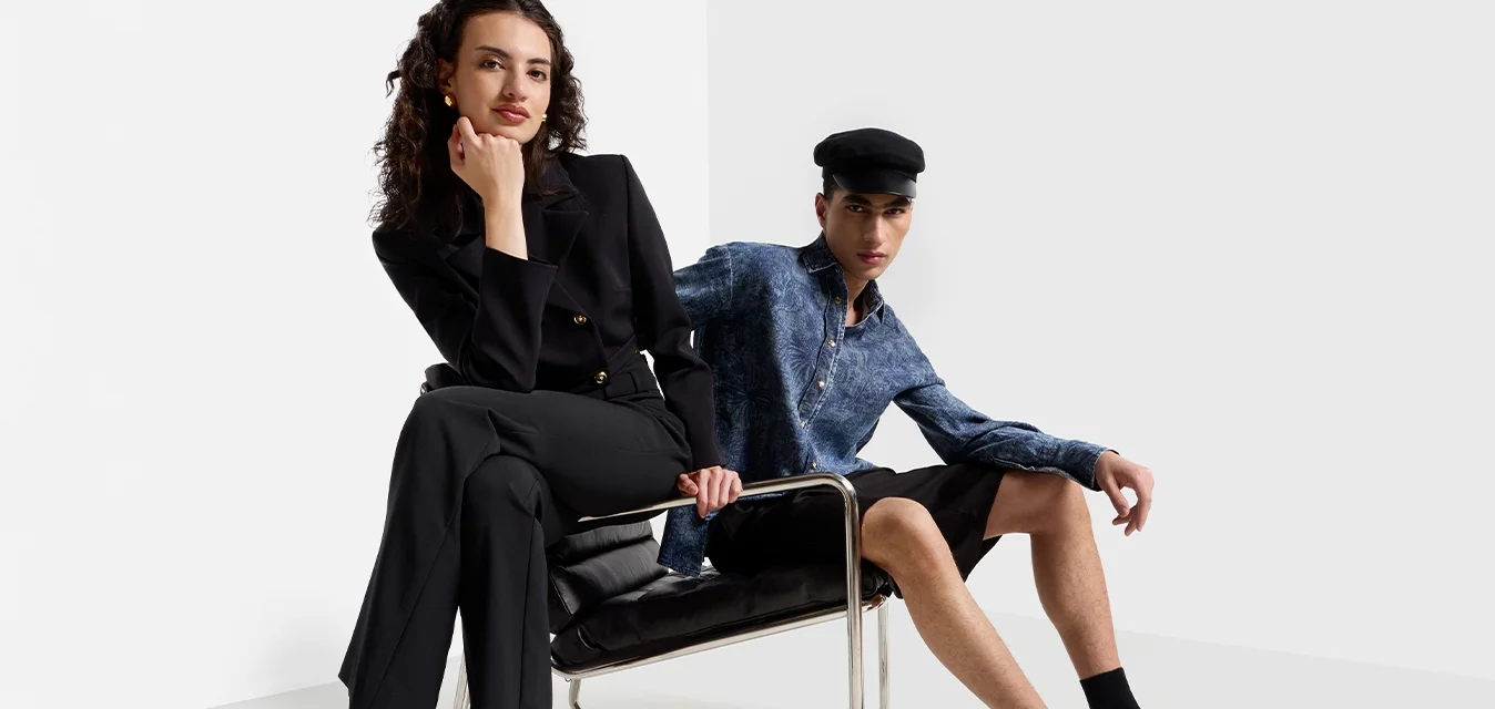  due modelle posano in un ambiente minimalista. Una indossa un completo nero e tacchi neri con cinturini; l'altra sfoggia una camicia blu e pantaloni corti neri, seduta.