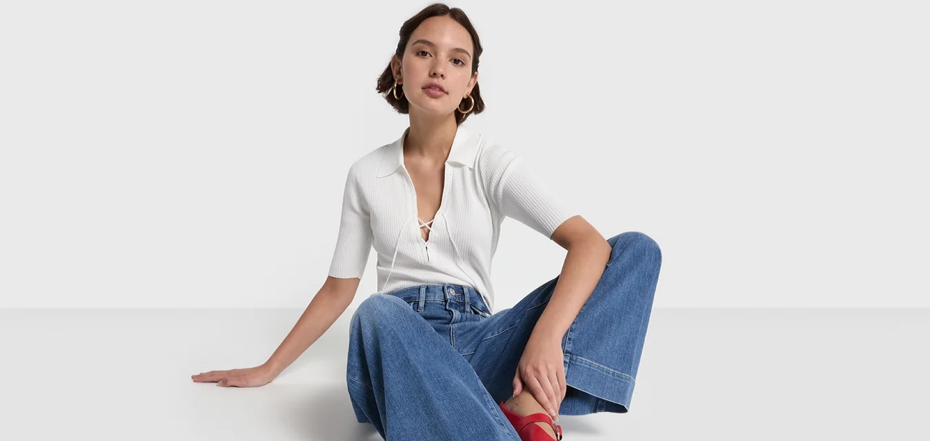 Blusa bianca a coste con maniche corte, abbinata a jeans larghi blu chiaro e scarpe con tacco quadrato rosse, seduta su uno sfondo semplice.