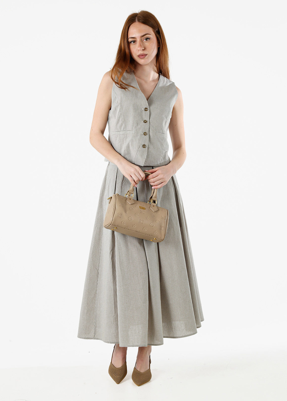Woman in grey gingham sleeveless vest and matching maxi skirt holding beige handbag, wearing brown pointed heels.