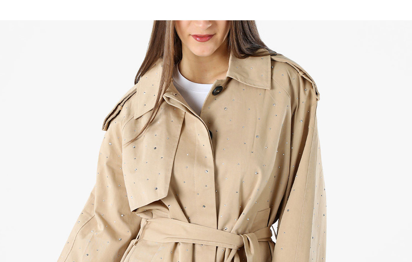 Model wearing beige belted trench coat with small crystal embellishments and white top on plain background