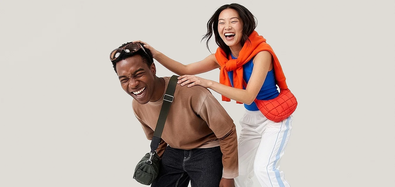 Giovane uomo che ride piegato in avanti con borsa a tracolla, donna dietro di lui sorridente, mani sulle sue spalle, indossa maglione arancione e sandali.