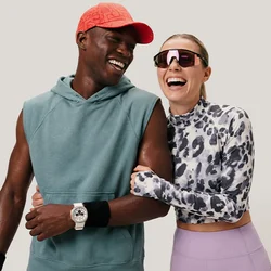 Uomo con felpa senza maniche e cappellino rosso sorride con donna con top fantasia e occhiali da sole mentre camminano braccio nella braccio su sfondo uniforme.