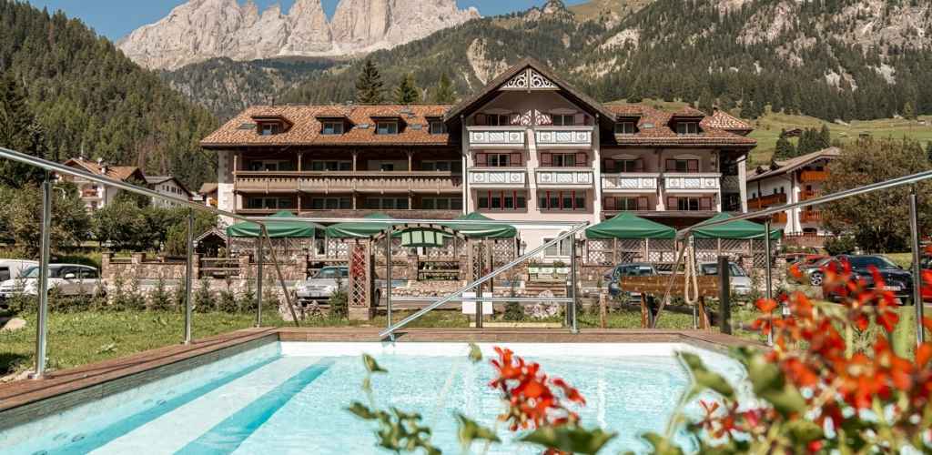 Outdoor swimming pool in front of alpine chalet hotel with red flowers, wooded hills and jagged peaks of the Dolomites