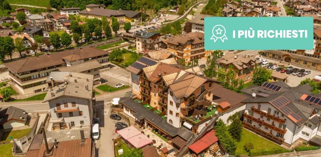 Aerial view of an alpine village with hotels and chalets among green hills, parking and a teal 'I PIÙ RICHIESTI' banner