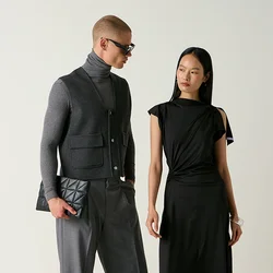Uomo con dolcevita grigio, gilet e pantaloni che tiene una pochette, e donna con abito nero e scarpe in piedi vicino a sedie, una delle quali è rovesciata sul pavimento.
