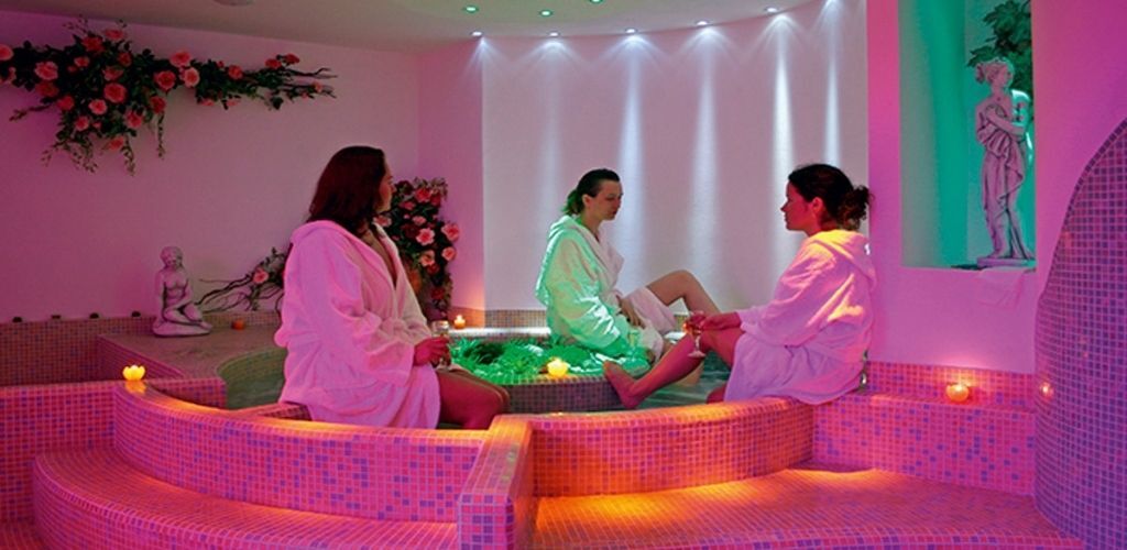 Three women in white robes relaxing in a pink-lit spa with mosaic seating, candles and decorative statues