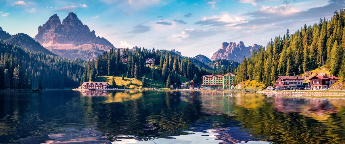 Alpine mountain lake with pine-covered slopes, lakeside hotels and rugged peaks reflected in calm water at sunset