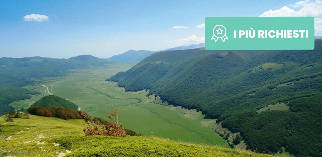 Panoramic green valley and forested hills under a blue sky with a teal 'I PIÙ RICHIESTI' badge in the top-right corner