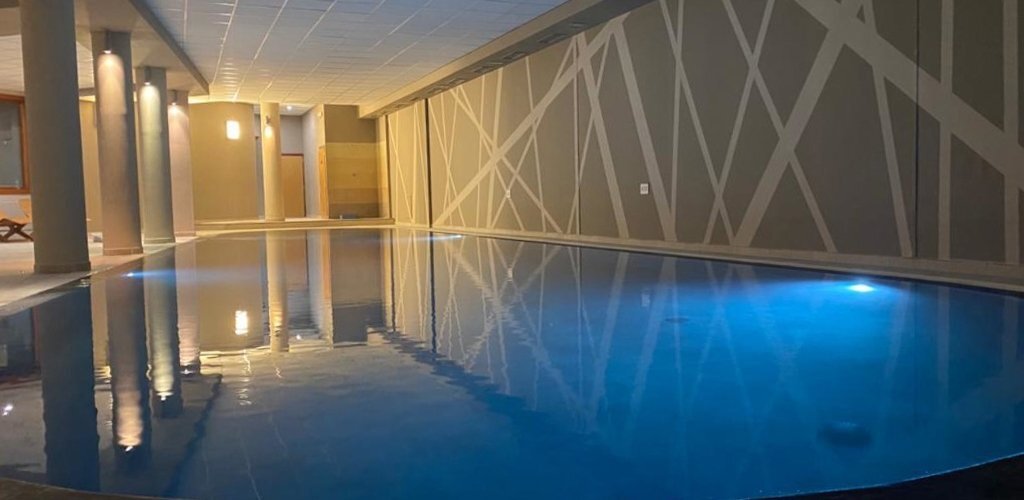 Modern indoor pool with calm blue-lit water, geometric patterned wall, columns reflected in the surface and poolside loungers