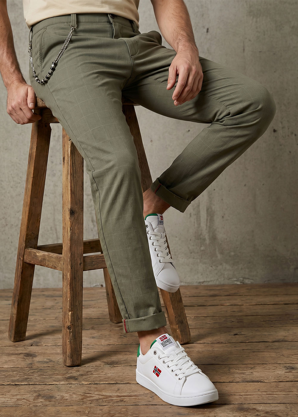 Man sitting on wooden stool wearing olive chinos with cuffed hems, chain accessory and white trainers with flag logo on wooden floor