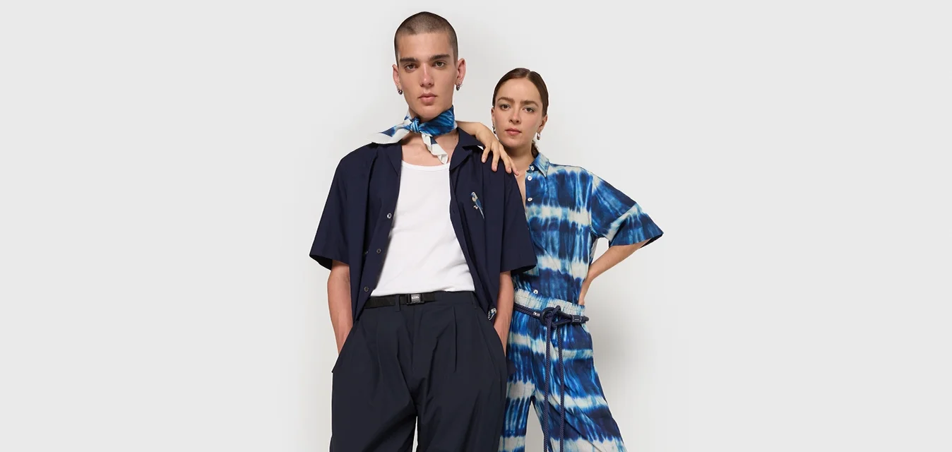 Due modelli posano insieme. Uno indossa una camicia blu scuro e pantaloni larghi; l'altro ha una tuta tie-dye blu. Entrambi hanno accessori minimal.