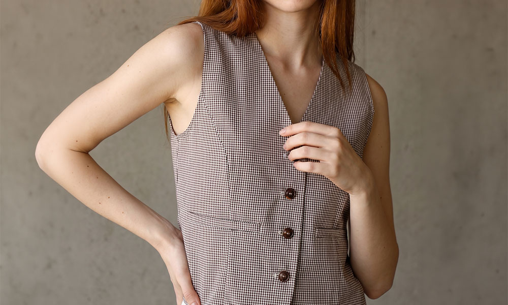 Close-up of woman wearing a sleeveless brown checked waistcoat, hand on chest and hip against a plain concrete background