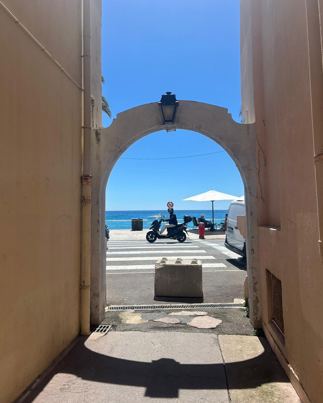 Archway framing a sunlit seafront street with a scooter at a zebra crossing, white parasol and blue sea