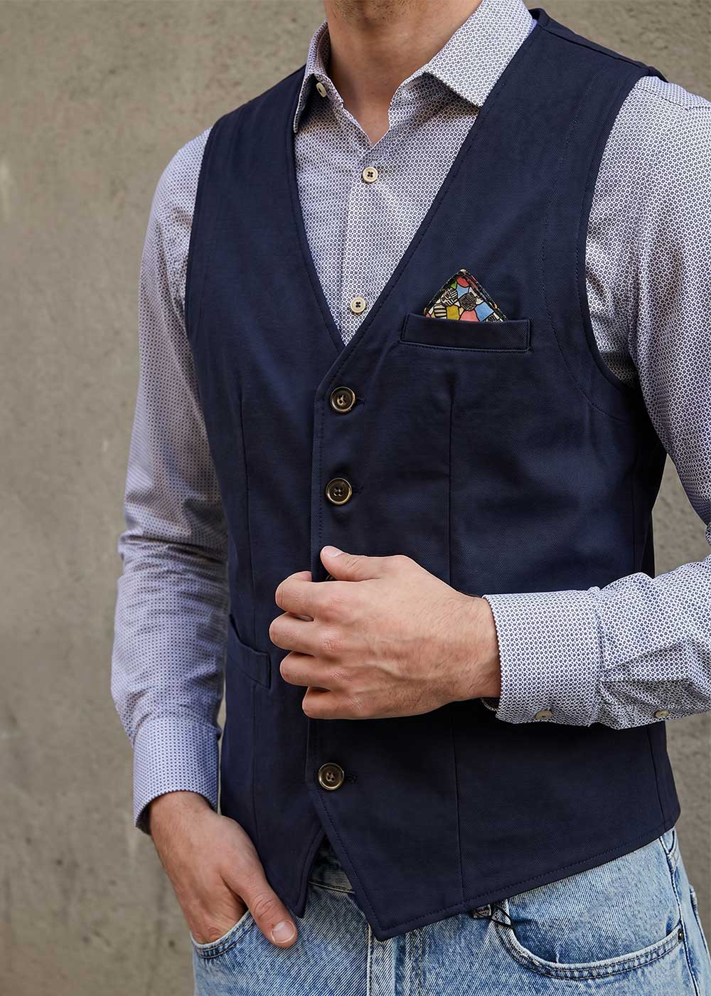 Man wearing a navy waistcoat over a patterned shirt with colourful pocket square, hand adjusting waistcoat while wearing casual jeans