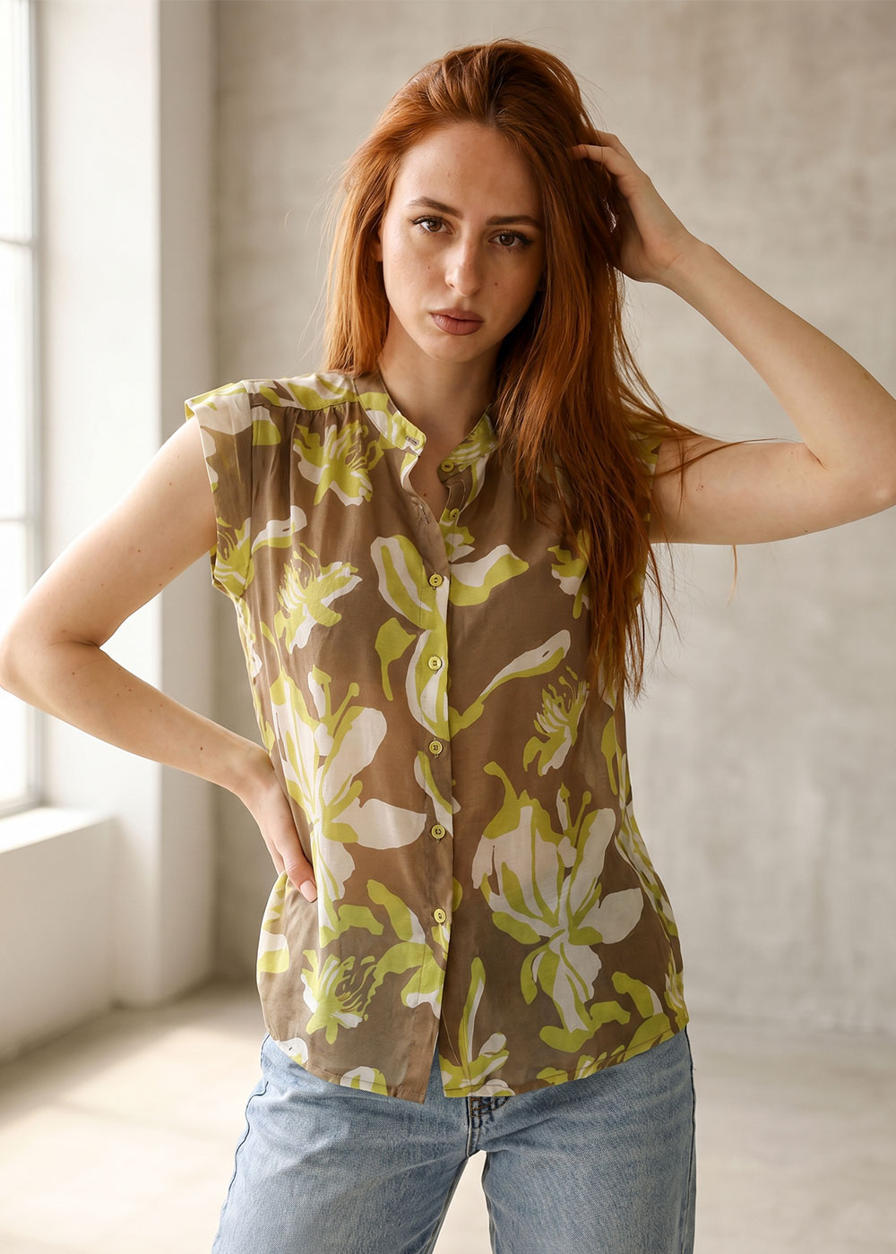 Red-haired woman in a green floral blouse and light blue jeans, standing by a window with one hand in her hair