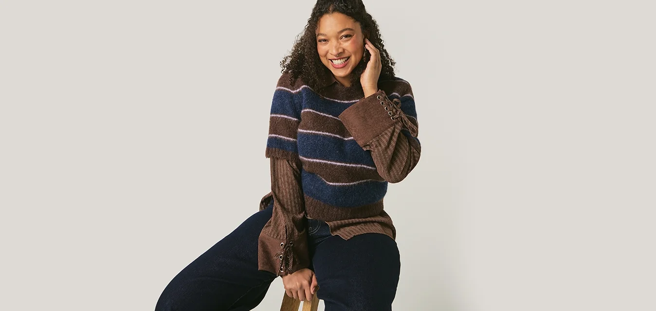 Giovane donna con capelli ricci, indossa un maglione a righe e pantaloni larghi, seduta su uno sgabello, sorridente contro uno sfondo chiaro uniforme.