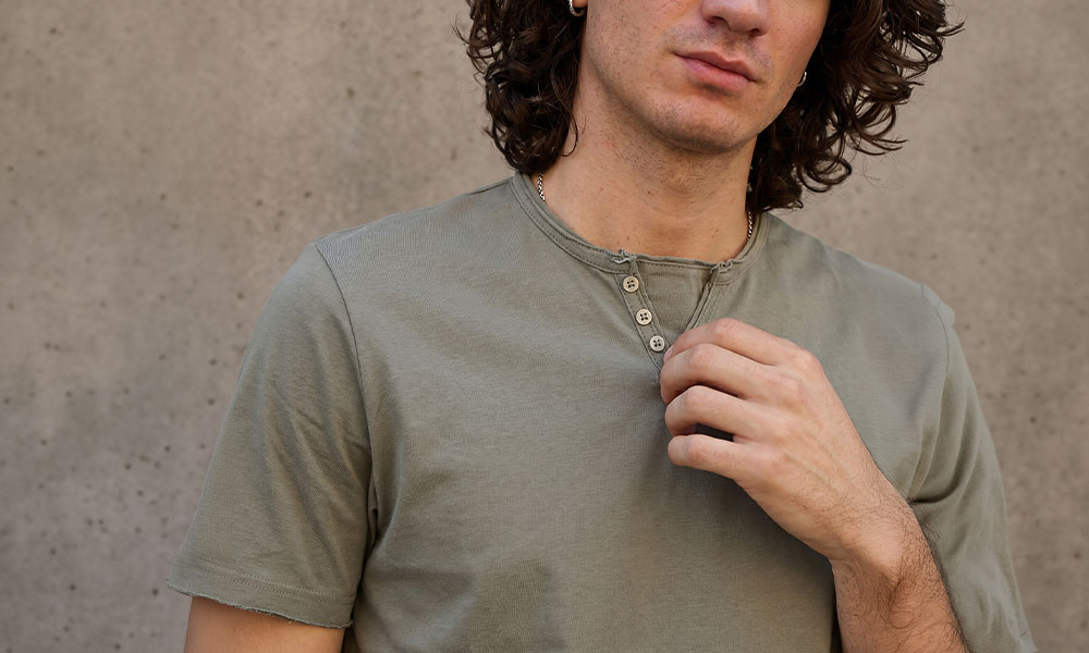 Young man adjusting buttoned olive-green henley T‑shirt in a cropped portrait against a textured concrete wall