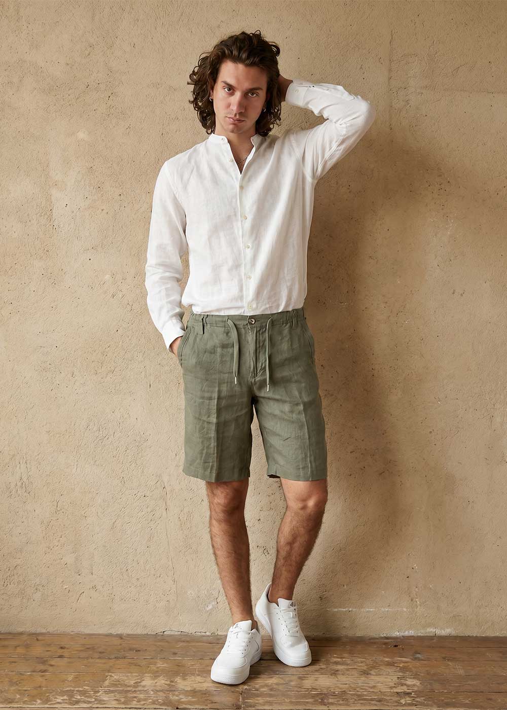 Man wearing white linen shirt, olive linen drawstring shorts and white trainers, standing against a textured wall
