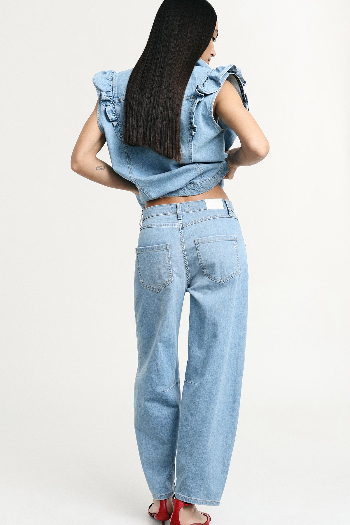 Rear view of model in light-blue denim vest and wide-leg jeans, long straight hair and red heels on pale background