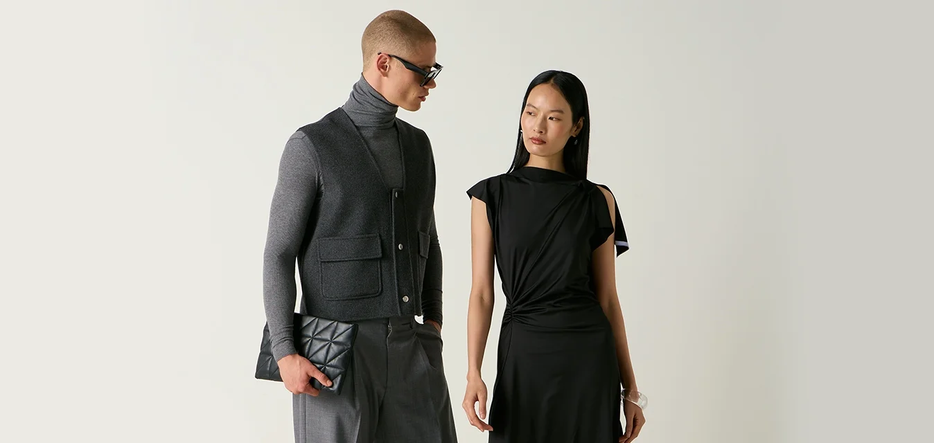 Uomo con dolcevita grigio, gilet e pantaloni che tiene una pochette, e donna con abito nero e scarpe in piedi vicino a sedie, una delle quali è rovesciata sul pavimento.
