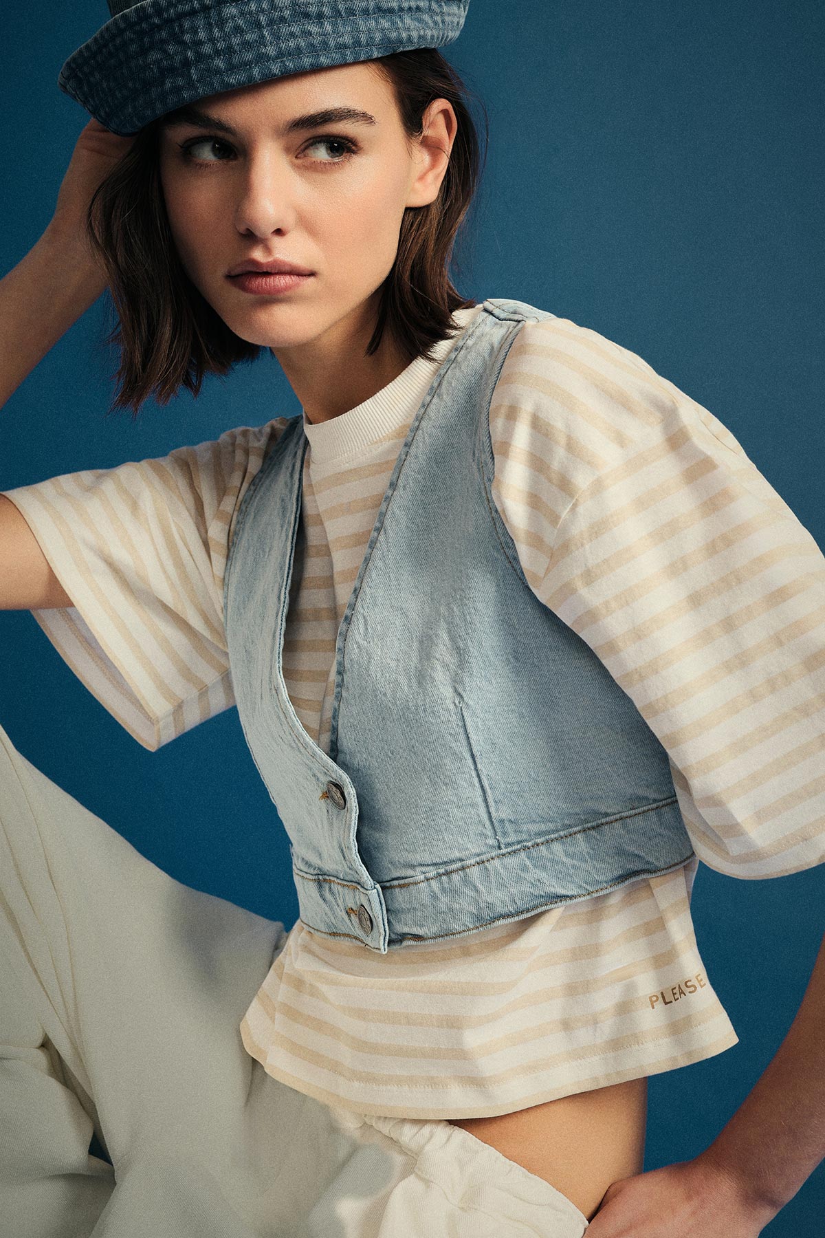Woman in blue bucket hat, striped cream tee and cropped denim vest against blue background, looking left