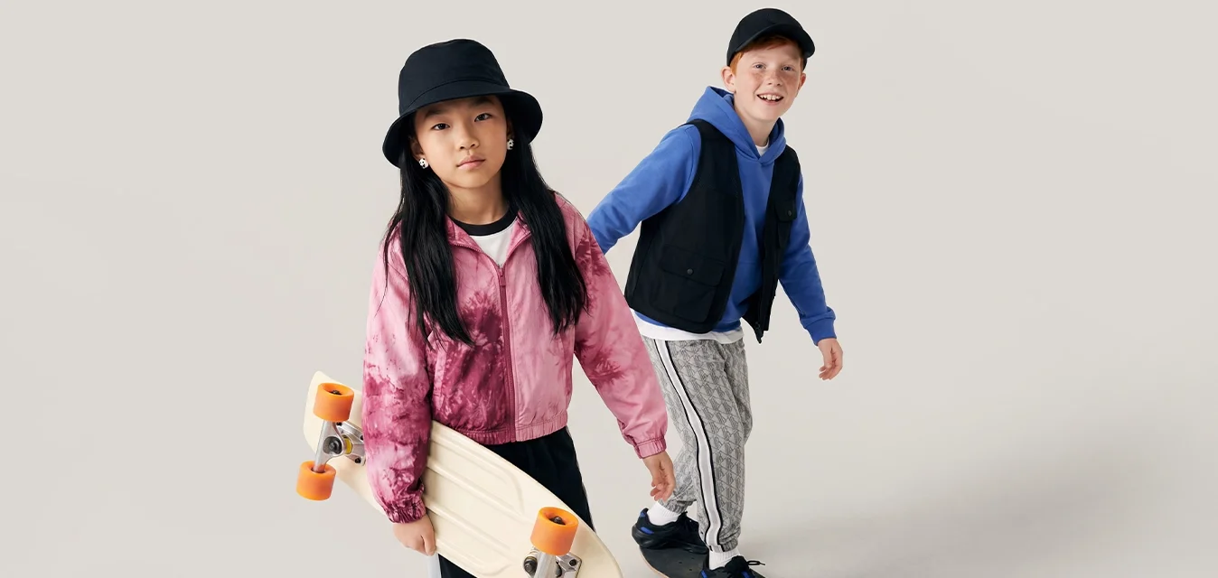 Due bambini fanno skateboard; la bambina tiene lo skateboard, il bambino cavalca la tavola, entrambi indossano abiti sportivi casual su uno sfondo chiaro e semplice.