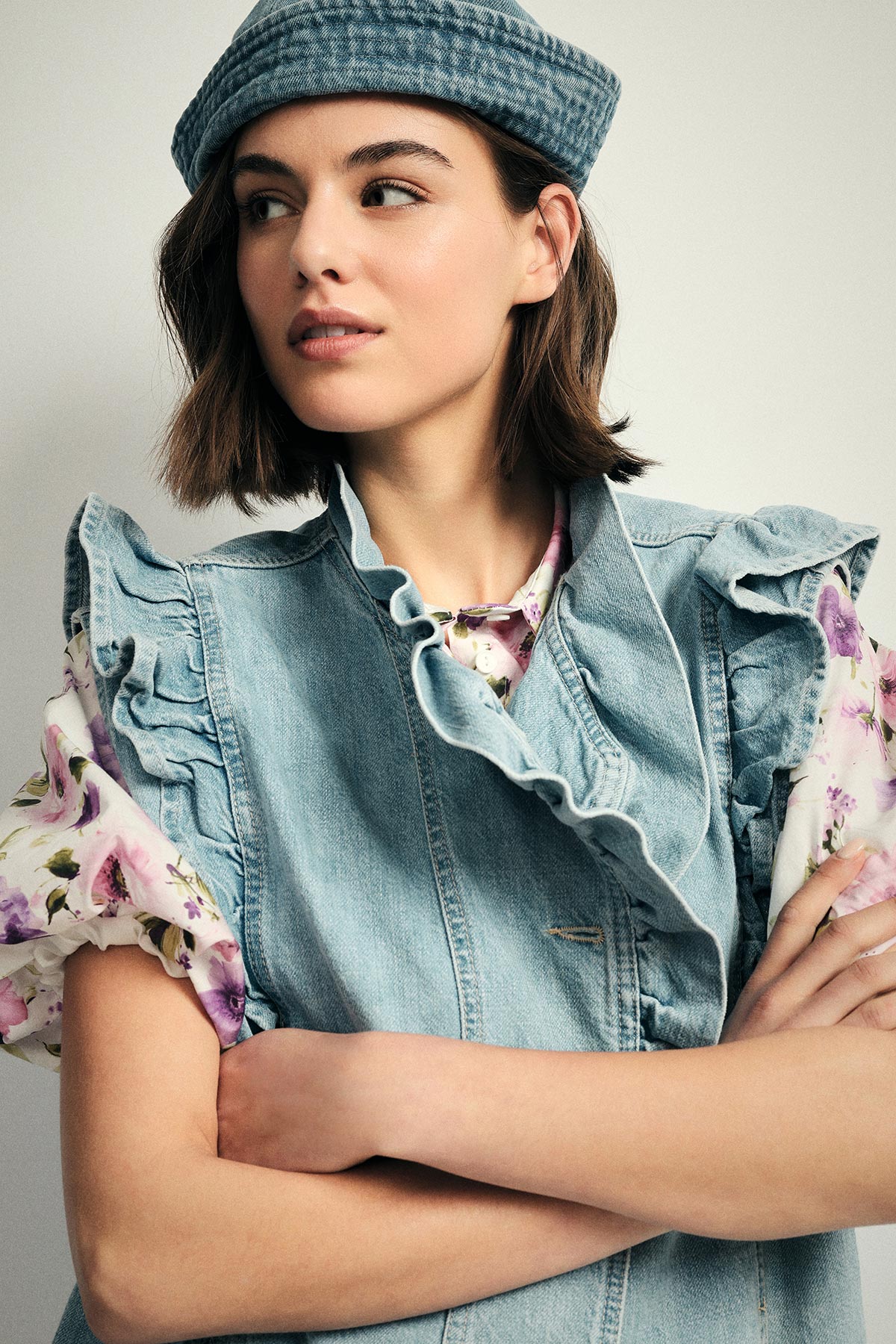 Young model wearing a denim ruffled vest and floral blouse with a matching denim cap, arms crossed and looking to the side