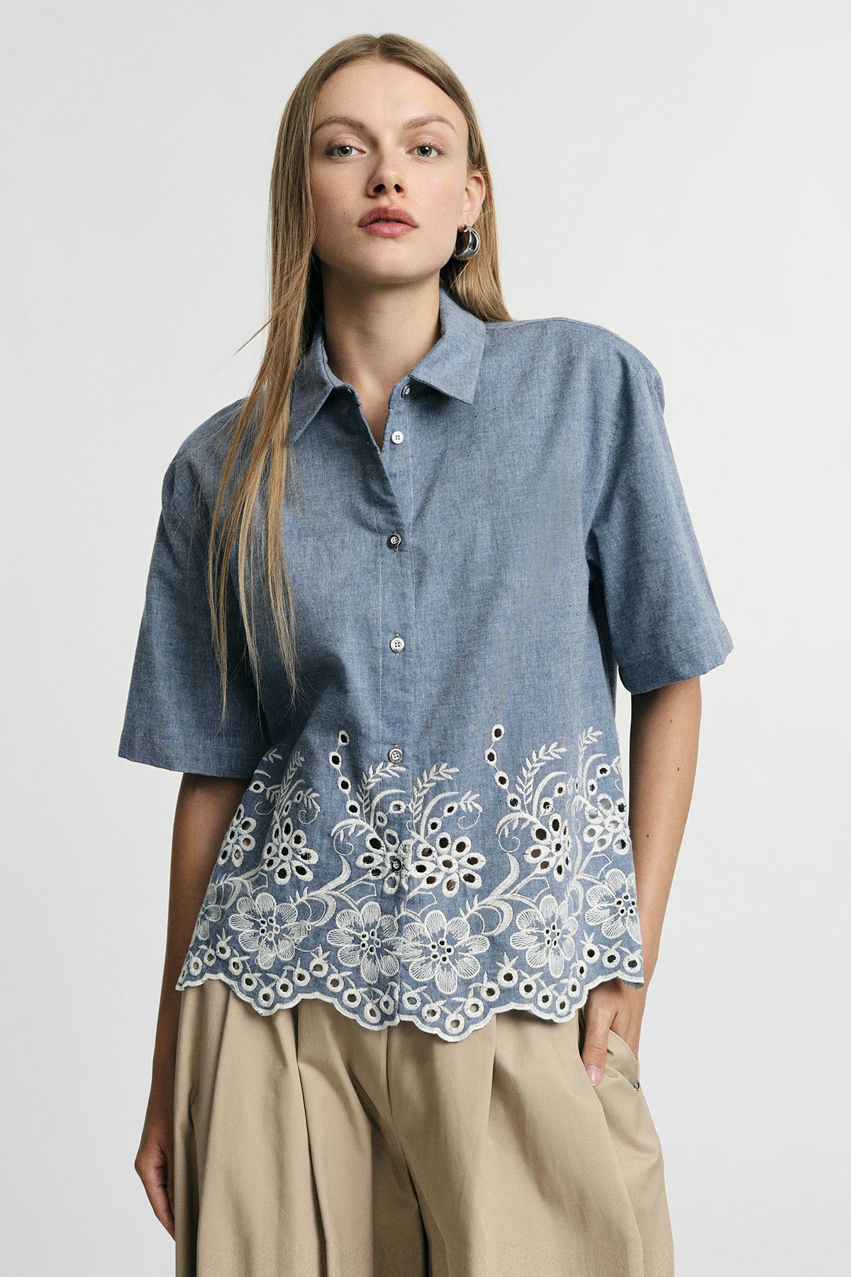Young woman wearing a short-sleeve blue chambray blouse with white floral eyelet embroidery and scalloped hem, paired with beige trousers
