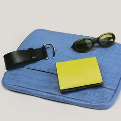 Clutch blu testurizzato con cintura in pelle nera, portafoglio giallo e occhiali da sole ovali scuri disposti su uno sfondo chiaro.