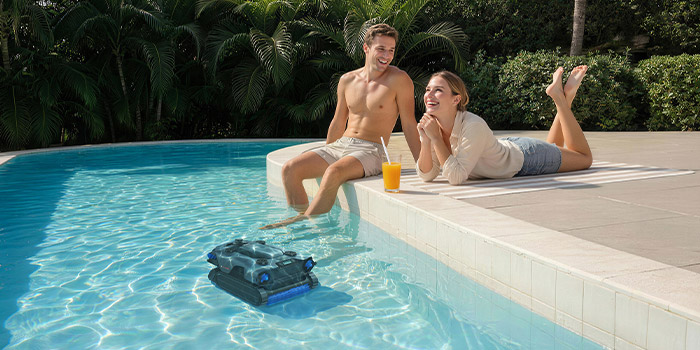 Due persone a bordo piscina che si rilassano mentre il robot pulisce la piscina