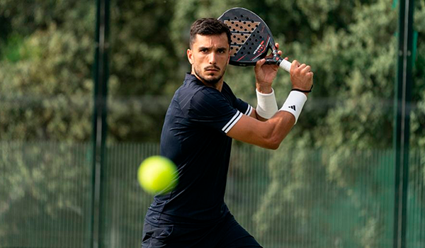 Padel player in black kit preparing a backhand shot on an outdoor court with ball in foreground