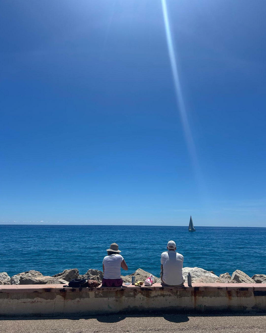 Two people sitting on a sea wall facing a deep blue sea and distant sailboat under a clear sky and sunbeam