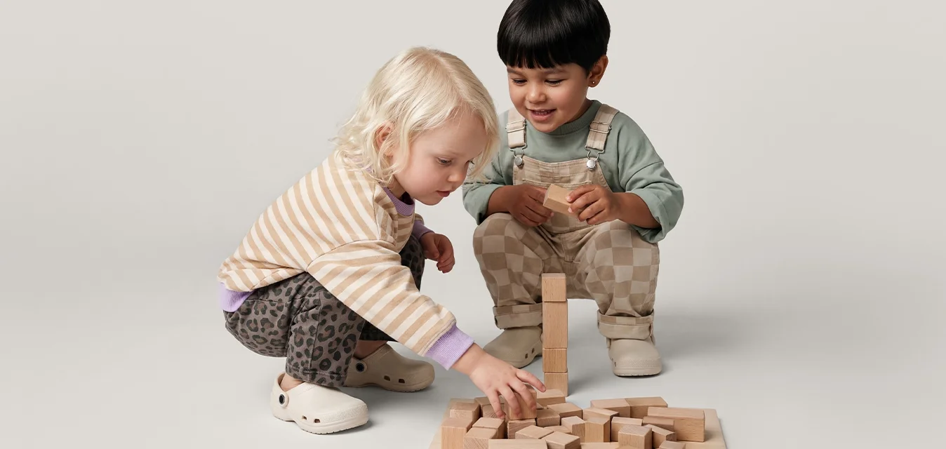 Due bambini piccoli seduti sul pavimento, che giocano con blocchi di legno disposti su una tavola piatta contro uno sfondo semplice.