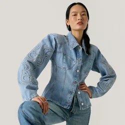 Donna con capelli lunghi e scuri che indossa una giacca di jeans azzurro chiaro ricamata e jeans abbinati, posando con un ginocchio sollevato davanti a uno sfondo semplice.