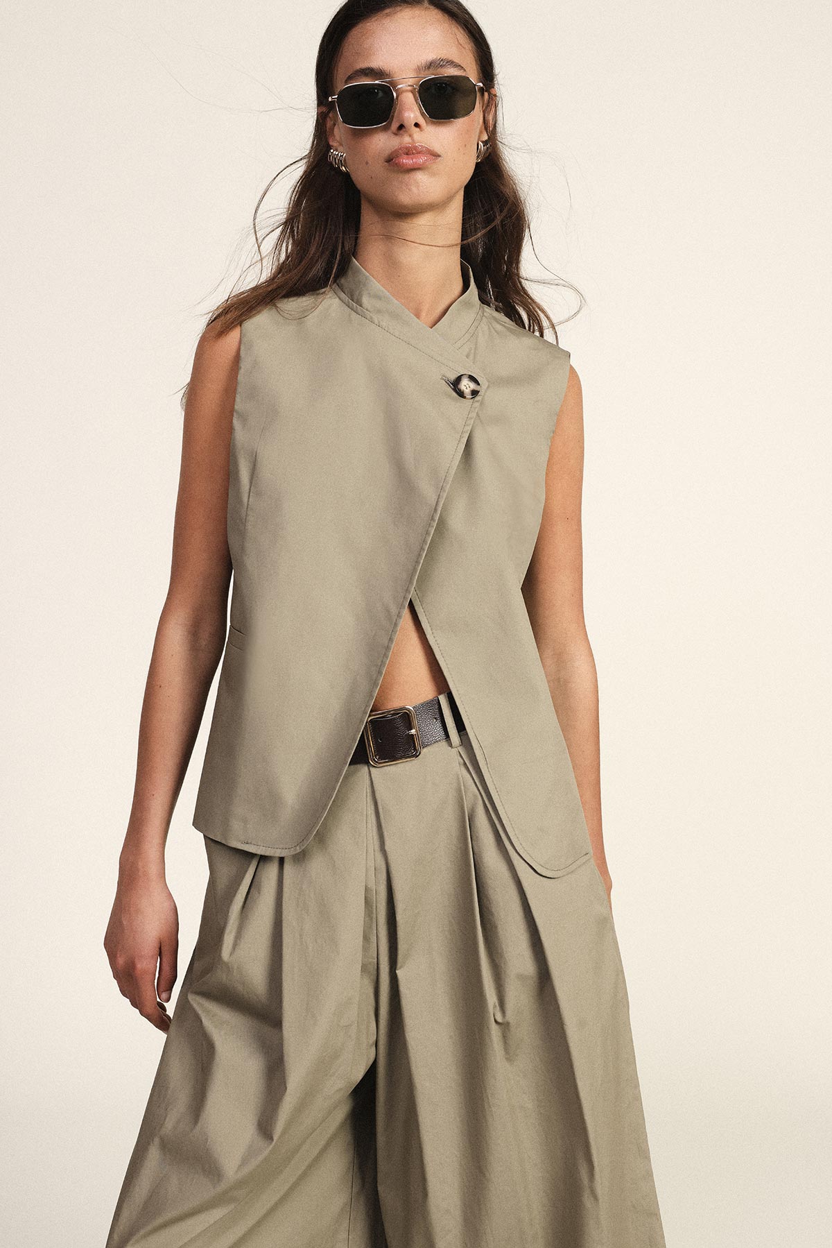 Woman wearing sleeveless beige wrap vest and matching wide-leg trousers with belt and sunglasses, studio fashion shot