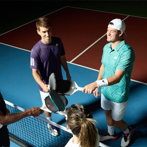 Four tennis players smiling and exchanging paddles at the net on an indoor hardcourt, preparing to start a match