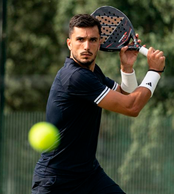 Male padel player in black kit preparing a backhand with paddle as a bright ball flies toward the camera on an outdoor court