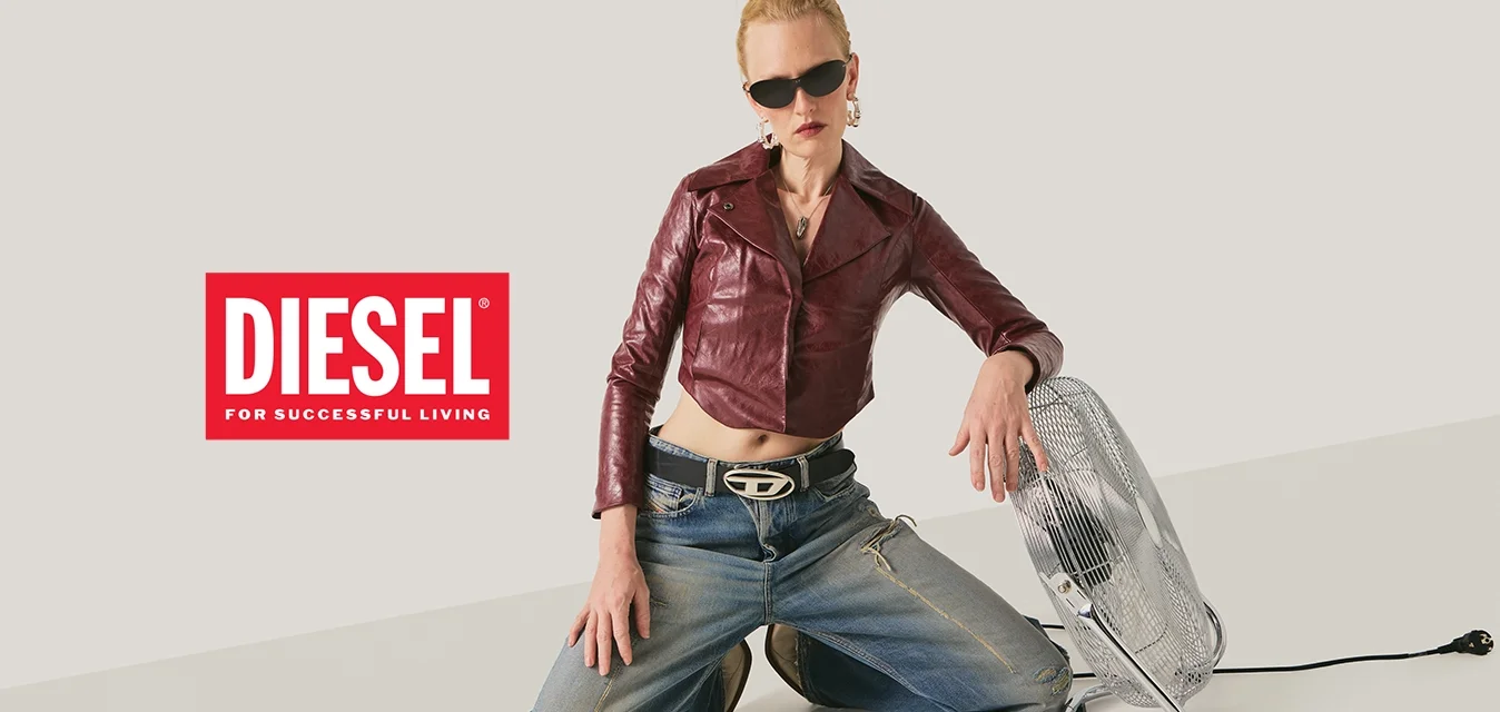 Persona in ginocchio sul pavimento che indossa occhiali da sole, una giacca di pelle color marrone scuro e jeans, con una mano appoggiata su un ventilatore da pavimento color argento.