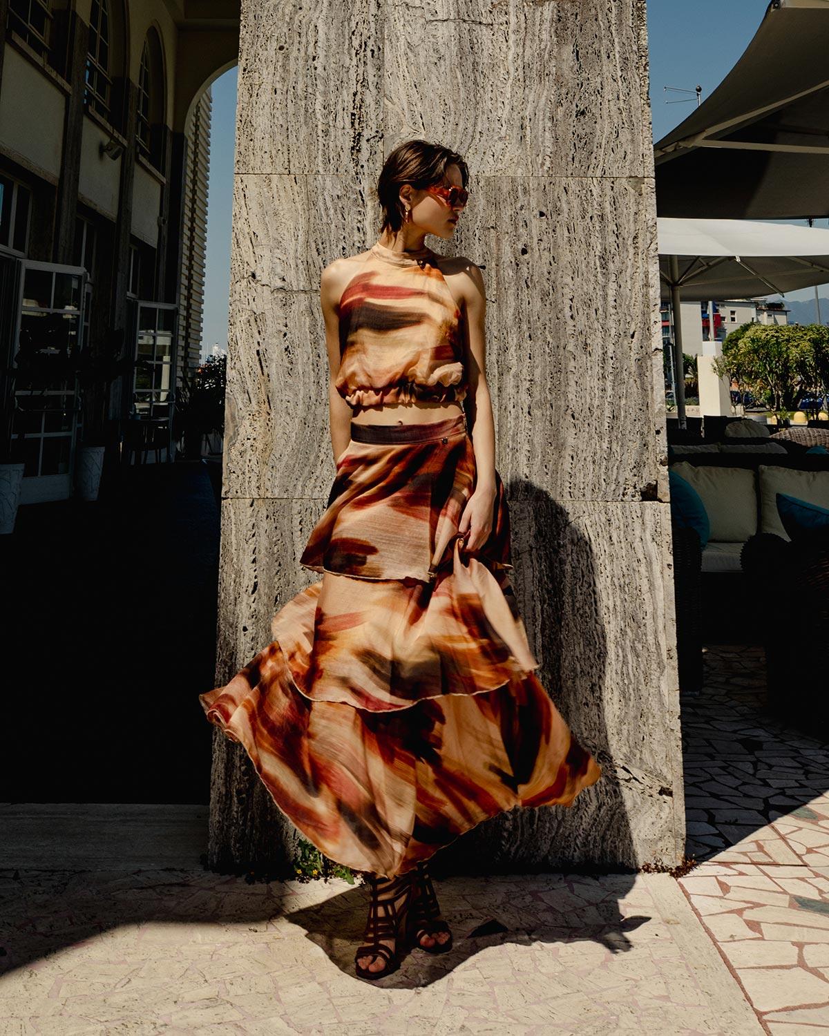 Woman in flowing brown and orange patterned summer dress and sunglasses leaning against a stone wall in a sunny outdoor plaza