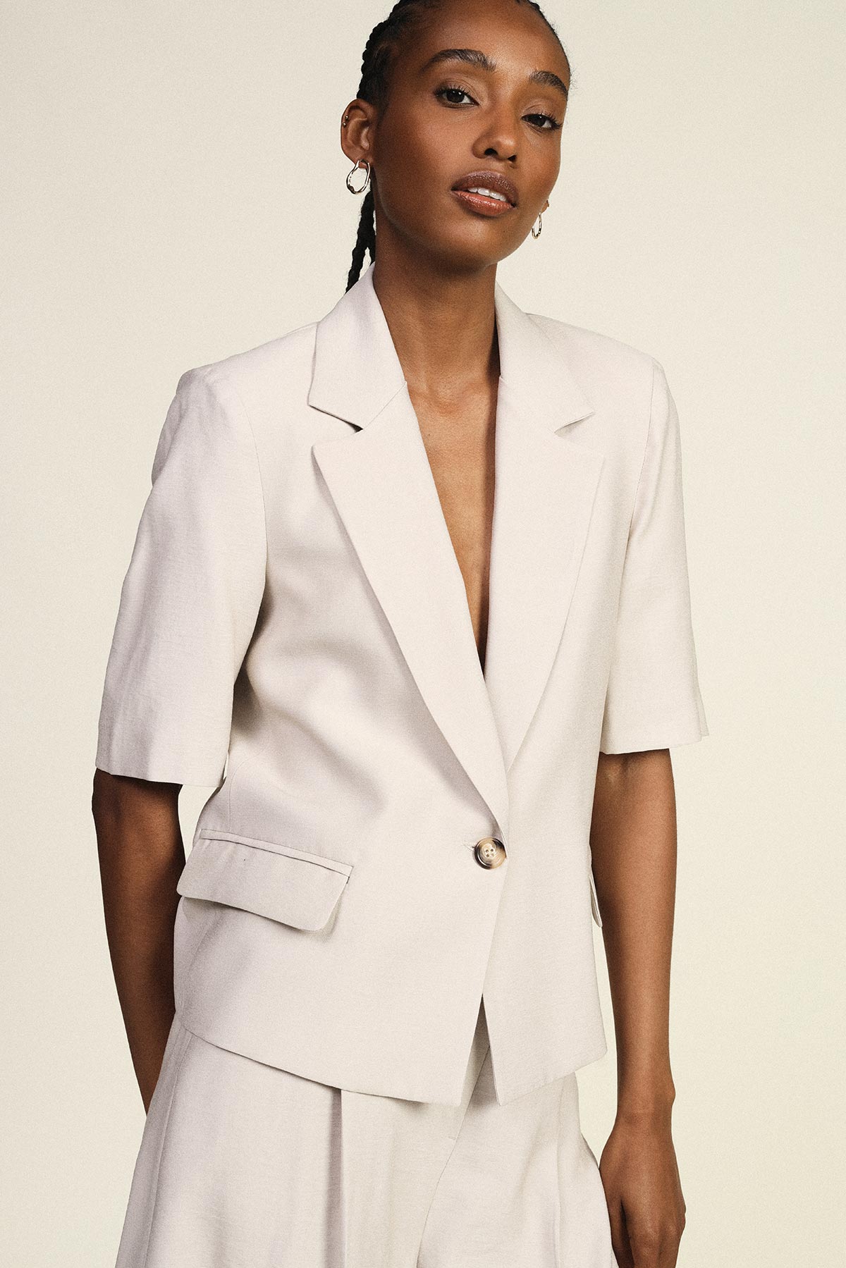 Woman in cream short-sleeve blazer and matching trousers, single-button tailored suit, hoop earrings, neutral studio background