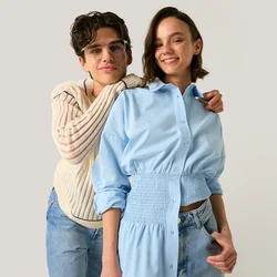 Due giovani donne stanno vicine, una con occhiali e maglione crema abbraccia un'altra con un vestito camicia blu, entrambe sorridenti su uno sfondo semplice.