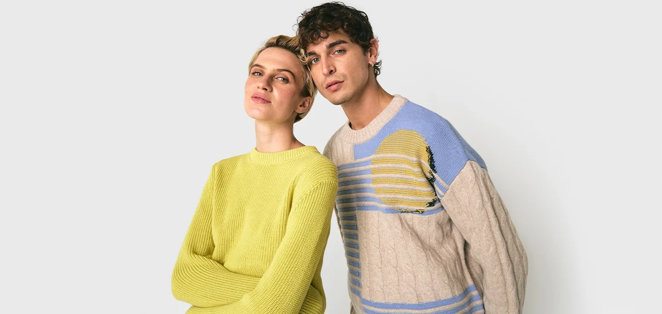 Due giovani adulti, uno con un maglione giallo e l'altro con un maglione beige e blu a fantasia, in piedi ravvicinati su uno sfondo bianco.